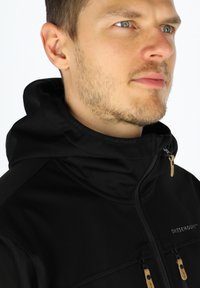 Black softshell jacket with a high collar, featuring two front pockets, zippers, and leather accents. Smooth texture with a modern design.