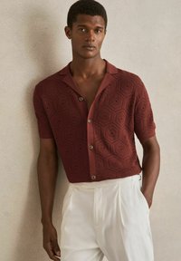 Cămașă tricotată cu mânecă scurtă, de culoare burgundă, cu o textură patterned ce prezintă forme hexagonale, guler cu nasturi și nasturi contrastanți.