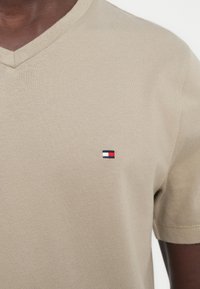 Μπεζ T-shirt με V λαιμόκοψη, φορεμένο από ένα άτομο, με μικρό ορθογώνιο λογότυπο σε ναυτικό, λευκό και κόκκινο χρώμα στο αριστερό στήθος.