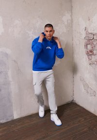 Modra kapuca s logotipom "YOUTH", sivi trenirki s stranskimi žepi in beli športni čevlji z modrimi detajli. Stoji ob teksturirani steni.
