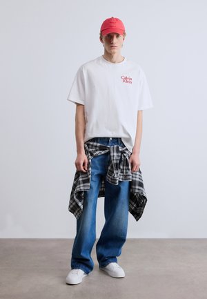 Biele bavlnené tričko s červeným logom "Calvin Klein", modré džínsy z denimu, čierno-biela kockovaná košeľa previazaná v páse, ružová basebalová čiapka, biele tenisky.