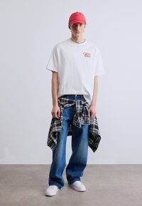 Λευκό βαμβακερό t-shirt με κόκκινο λογότυπο "Calvin Klein", μπλε τζιν παντελόνι, ασπρόμαυρο καρό πουκάμισο δεμένο στη μέση, ροζ μπέιζμπολ καπέλο, λευκά αθλητικά παπούτσια.