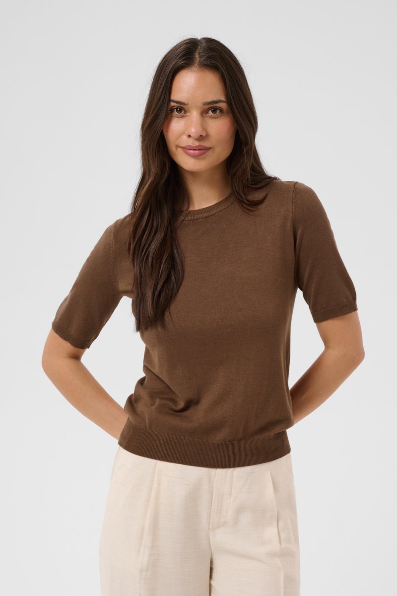 Femme aux longs cheveux foncés portant un pull marron à manches courtes et un pantalon beige clair, debout les mains dans le dos.