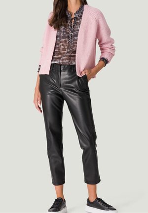 Mujer con un cárdigan de punto rosa sobre una blusa estampada, pantalones de cuero negros recortados y zapatillas negras con suelas blancas.