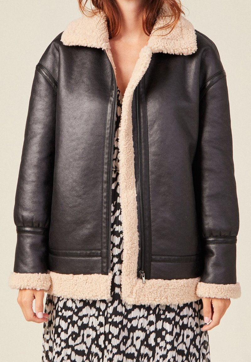 Woman wearing a black leather jacket with beige shearling lining and cuffs over a black and white patterned dress.