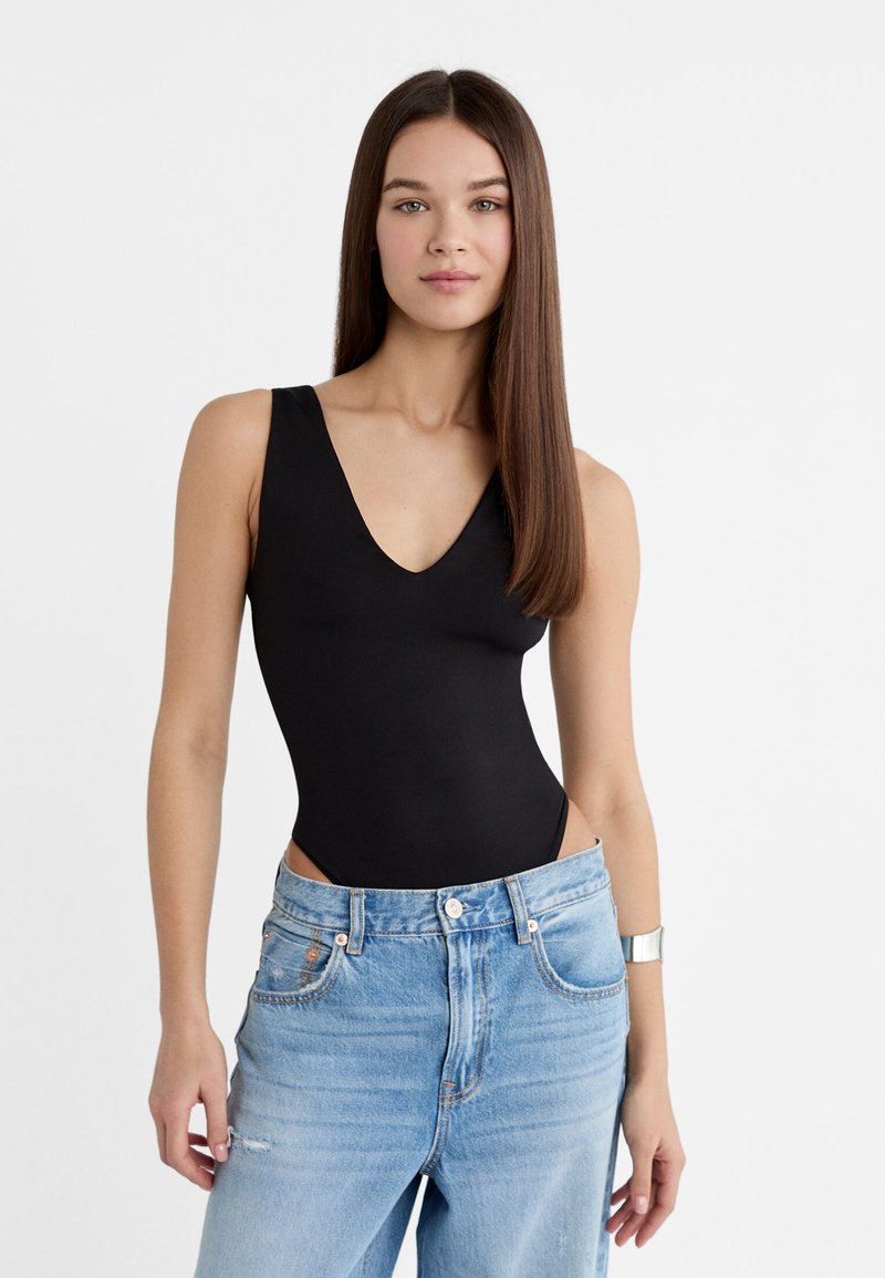 Zwarte bodysuit met een diepe V-hals en brede banden, gecombineerd met lichtblauwe high-waisted jeans, met lichte slijtage en een klassieke snit.