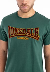 Grøn bomulds T-shirt med "LONSDALE LONDON" logo i gult og burgunder. Rund hals og korte ærmer, glat tekstur.