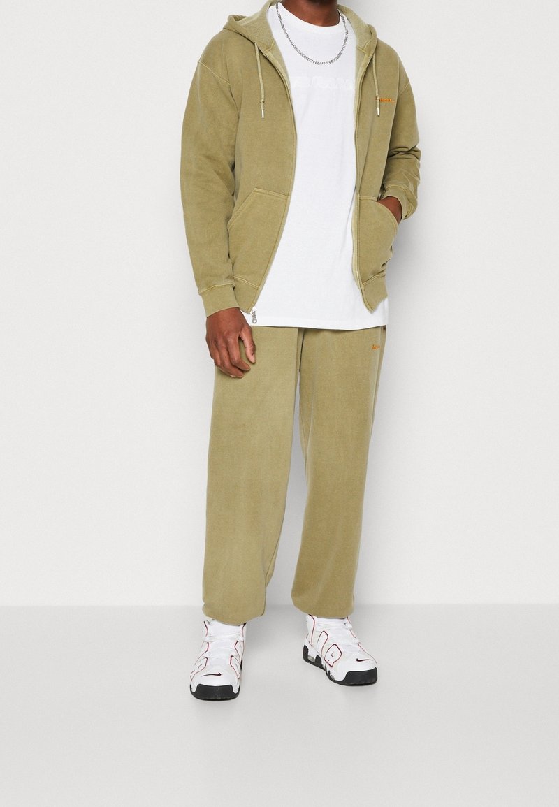 Homme portant un sweat à capuche vert olive et un pantalon assorti, un t-shirt blanc, un collier chaîne argentée et des baskets blanches avec semelles noires et accents rouges.