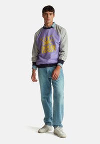 Felpa viola con maniche grigie con grafica "Stranger Things" in giallo. Indossata con jeans azzurri e scarpe da ginnastica bianche.