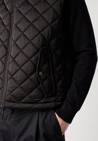 Black quilted vest with a high collar, front zipper, and side pocket. The garment features a smooth texture and is layered over a black sweater.