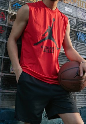 Mann in rotem ärmellosem Chicago Bulls Shirt und schwarzer Shorts, der einen Basketball hält und vor einer mit Graffiti bedeckten Glassteinwand steht.