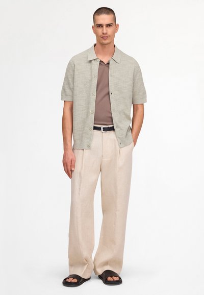 Uomo con capelli corti indossa una camicia grigio chiaro a maniche corte con bottoni sopra una maglietta marrone, pantaloni beige a gamba larga, cintura nera e sandali neri con punta aperta.