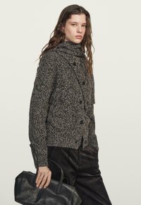 Cardigan lavorato a maglia di colore grigio scuro con collo alto, caratterizzato da bottoni sul davanti. Abbinato a pantaloni in velluto a coste neri e una borsa in pelle.