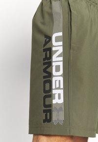 Olivgröna shorts med elastisk midja, med en vertikal vit och svart "UNDER ARMOUR" grafisk design på sidan. Slät textur.
