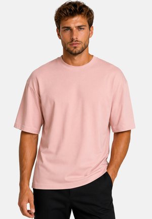 Uomo con capelli corti e ondulati che indossa una maglietta rosa chiaro larga e pantaloni neri, in piedi con una mano in tasca su sfondo bianco.