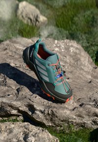 Shoe de hiking azul e preto com atacadores laranja e solas repousando sobre uma superfície rochosa com relva verde ao fundo.