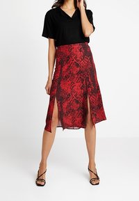 Röd ormtryckt kjol med en hög slitsdesign, tillverkad av lättviktsmaterial. Kombinerad med en svart t-shirt med kort ärm och svarta sandaler med remmar.