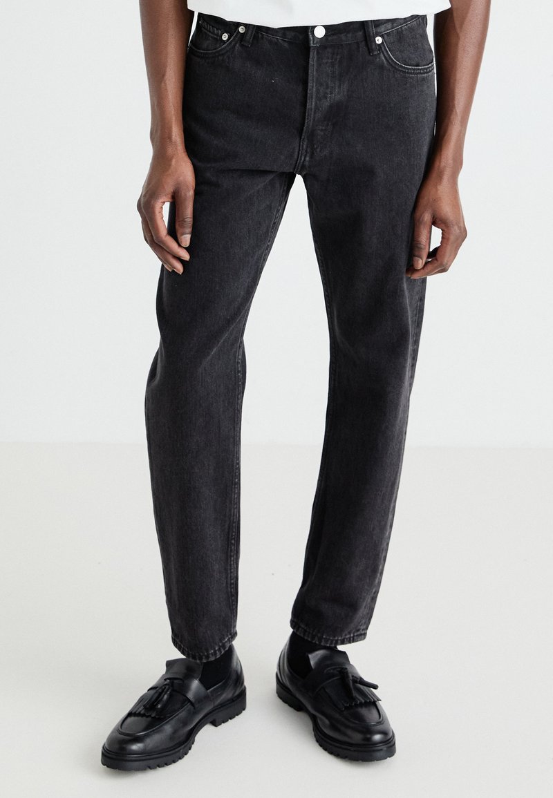 Jean noir en denim à coupe droite, avec un design à cinq poches et une légère décoloration. Porté avec des mocassins noirs et des chaussettes noires.
