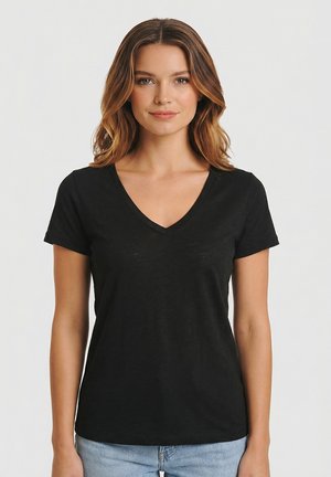 Femme aux cheveux bruns ondulés portant un T-shirt noir à manches courtes avec col en V et un jean bleu clair, sur un fond clair uni.