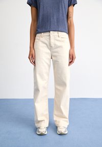 Jean droit couleur crème, taille haute, avec fermeture à boutons et poches avant, associé à un T-shirt bleu marine et des baskets grises.