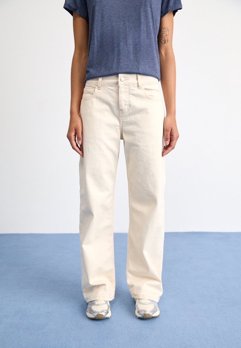 Jean droit couleur crème, taille haute, avec fermeture à boutons et poches avant, associé à un T-shirt bleu marine et des baskets grises.