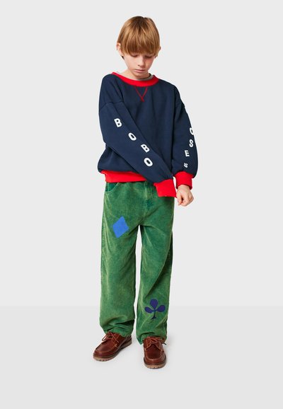 Sweatshirt blu scuro con dettagli rossi e testo bianco, abbinato a pantaloni in velluto a coste verdi con toppe e forme blu. Scarpe marroni con lacci.