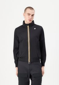 Čierna zippová bunda s vysokým golierom, s žltými a bielymi prúžkami na zips. Mäkko, hladká tkanina s jemným leskom.