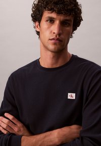 Mann mit lockigem dunklem Haar, trägt schwarzen Calvin Klein Pullover, Arme verschränkt, neutraler Gesichtsausdruck, einfarbiger hellgrauer Hintergrund.