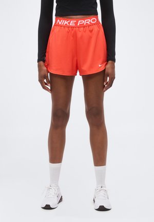 Personne portant un short de sport Nike Pro orange vif, des chaussettes blanches montant jusqu’à la cheville et des baskets Nike blanches sur un fond clair uni.