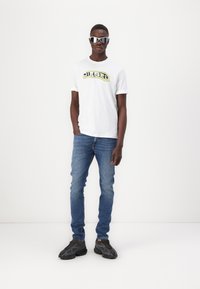 White cotton t-shirt with "DIESEL" logo in yellow and black, blue denim jeans, and black textured sneakers with chunky soles. Silver sunglasses.