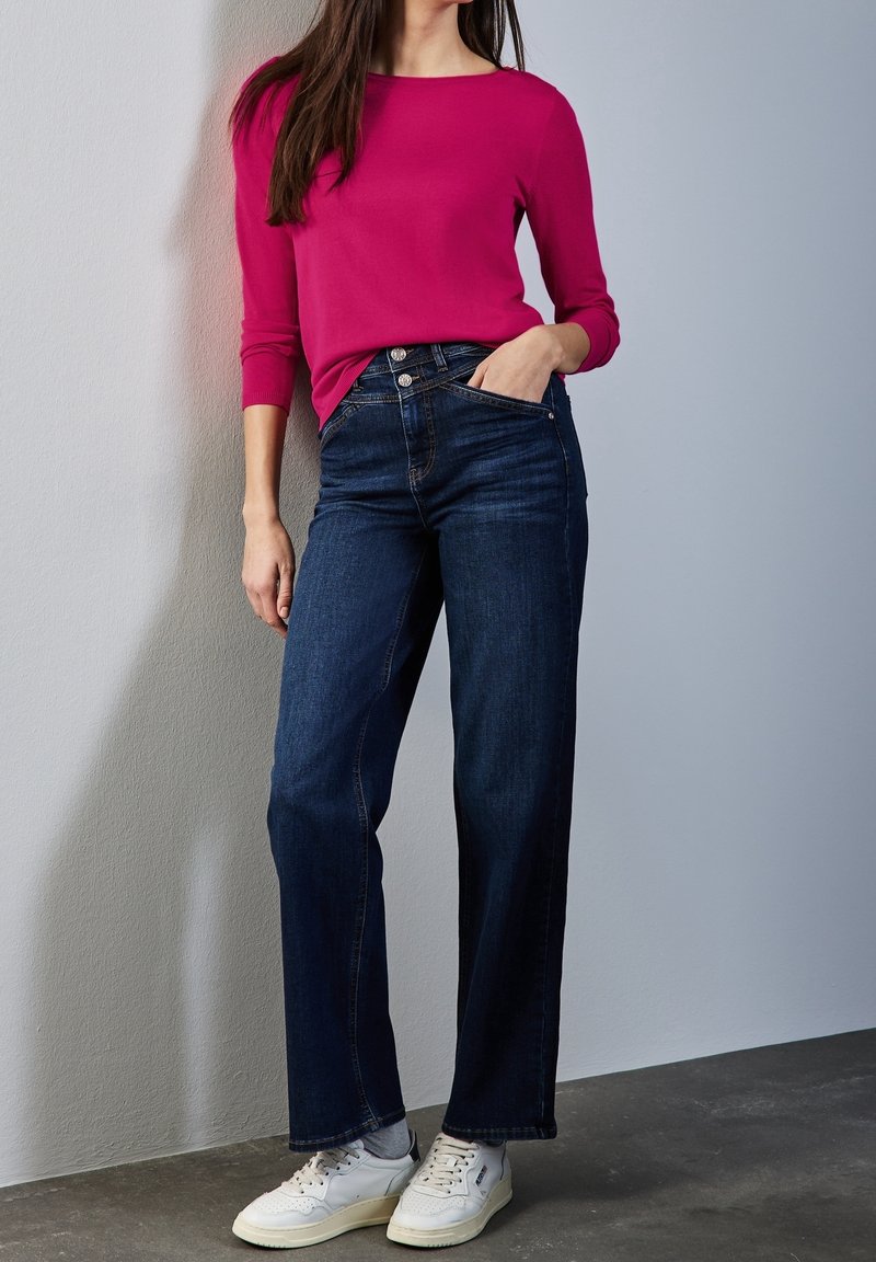 Femme portant un haut à manches longues rose vif, un jean taille haute bleu foncé, des chaussettes grises et des baskets blanches, debout contre un mur clair.