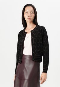 Cardigan noir avec un motif floral, bords côtelés et fermeture à boutons. Porté sur un haut clair et associé à une jupe en cuir bordeaux.