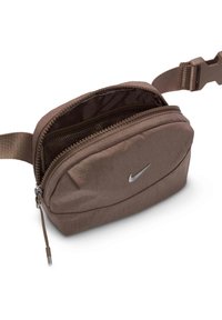 Hnedá textilná taška s zaobleným tvarom, strieborným zipsom a malým strieborným logom Nike. Pripojený nastaviteľný popruh na nosenie.