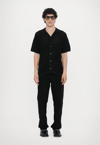 Juoda trumpų rankovių mygtukinė marškiniai su tekstūruotu audiniu ir džinsais, derinami su blizgiais juodais bateliais. Modelis dėvi didelius tonuotus akinius nuo saulės.