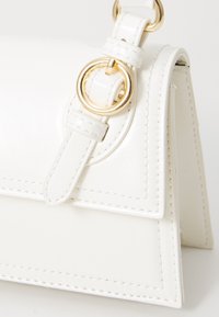 Sac à main en cuir blanc au design structuré, avec des accents en métal doré, une grande bague circulaire en évidence, et des détails de couture visibles.