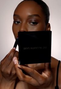 Compattino nero con scritte dorate tenuto vicino al viso, che mostra un prodotto per le labbra rosso. Carnagione e trucco sono naturali e mettono in risalto i lineamenti.