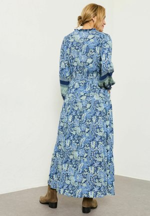 Frau mit blondem Haar trägt ein langes, blaues Blumenkleid mit Puffärmeln und braunen Stiefeletten, steht vor einer schlichten Wand.