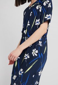 Laivansinis kukkainen mekko lyhyillä hihoilla, joustava vyötärö, jossa valkoisia kukkia ja vihreä/banaani-värisiä yksityiskohtia sujuvassa, kevyessä kankaassa.