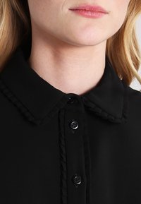 Černá blůza s volánkovým límcem, zapínáním na tlačítka s dvěma černými knoflíky a hladkým povrchem.