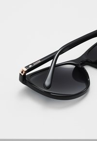 Ochelari de soare negri cu un cadru unghiular, supradimensionat. Prezintă o finisare lucioasă și un accent de aur roz pe braț, cu branding "FOSSIL".