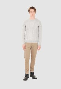Maglione grigio a trecce con polsini e orlo a coste, abbinato a chinos beige e stivali neri stringati, in piedi contro uno sfondo neutro.