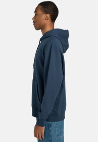 Navy blue hoodie made of cotton with a kangaroo pocket, drawstring hood, ribbed cuffs, and hem; worn with denim pants.