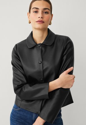 Woman wearing a black buttoned faux leather jacket with a rounded collar and blue jeans, arms crossed, neutral background.