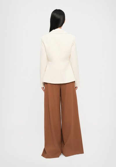Blazer de marfil a medida con hombros estructurados, combinado con pantalones marrones de pierna ancha. Textura de tela suave, líneas limpias y detalles visibles en la parte posterior.