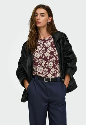 Femme portant une veste en cuir noire sur une chemise fleurie bordeaux, rentrée dans un pantalon bleu marine avec une ceinture noire, mains dans les poches.