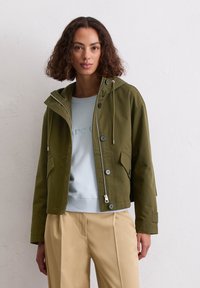 Przycięta oliwkowozielona kurtka z kapturem, z zamkiem błyskawicznym z przodu, metalowymi akcentami i zapięciami na guziki, w połączeniu z jasnoniebieskim swetrem i beżowymi spodniami.