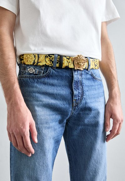 Schwarzer Gürtel mit goldener Löwenkopfschnalle, verziert mit einem gelben Blumenmuster. Über blauen Jeans getragen. Weißes T-Shirt sichtbar.