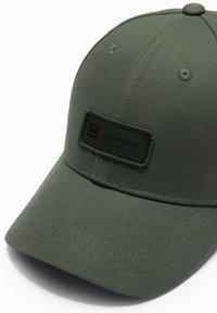 Grüne Baseballmütze aus Stoff mit gebogenem Schirm, versehen mit einem rechteckigen Logo-Patch in Schwarz und Rot. Sichtbare Nähte.