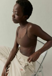 Vrouw in een bruine crop top en beige sweatpants zittend op een transparante stoel, met een bedachtzame blik opzij tegen een neutrale achtergrond.