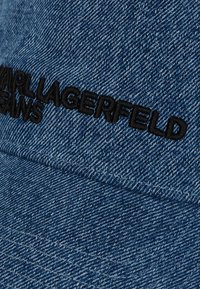 Denim cepure ar teksturētu zilo audumu, rotāta ar melnu izšuvumu "KARL LAGERFELD JEANS" uz priekšpuses.